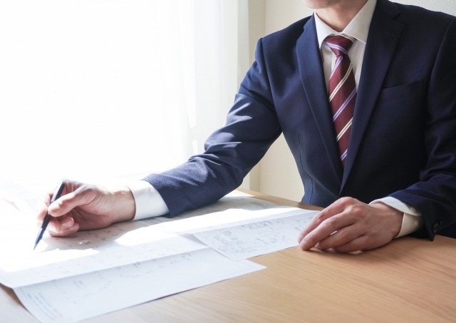 画像:男性のビジネスパーソンが資料をペンで指しながら説明をしている写真