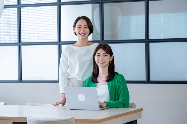 画像：パソコンを前に座る女性と後ろに立つ女性の写真